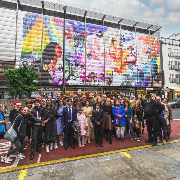 A diverse group of people stand smiling in front of a vibrant, colourful mural on a large building wall. The scene is lively and celebratory.