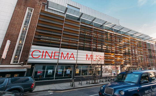 Modern cinema building with horizontal slats and large red letters reading "CINEMA" and "MUSIC." A blue taxi and a parked car are visible on the street.