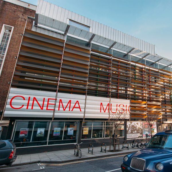 Modern cinema building with horizontal slats and large red letters reading "CINEMA" and "MUSIC." A blue taxi and a parked car are visible on the street.