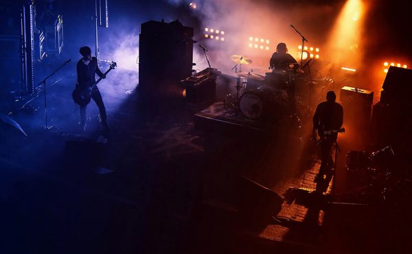 A three piece band playing music on stage. Half the stage lit with a blue light, the other orange. The bodies are silhouettes.