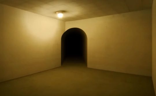 A vacant room with yellow walls. There is a dark tunnel ahead through an arched doorway. The space is dimly lit via a single, small lamp hanging from the ceiling.