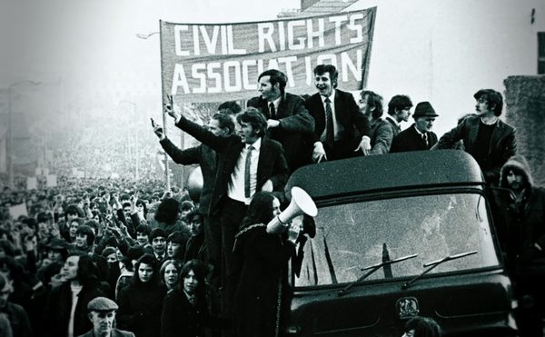 A black and white image. In the foreground, a truck carrying a group of protestors with the flag saying civil rights movement. Behind them a large crowd of protestors.