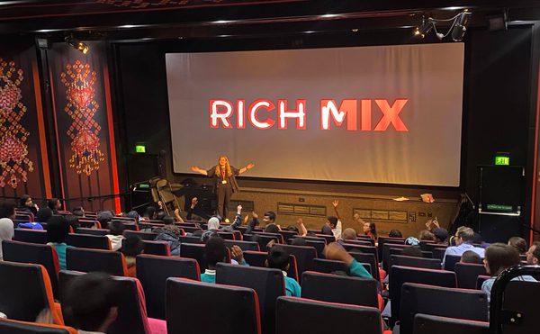 A cinema screen with the text Rich Mix on the screen. A bunch of children in seats have their arms raised, as does a person on a stage in front of the screen.