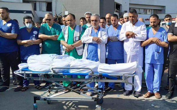 A group of Palestinian doctors stand solemnly behind a stretcher carrying a deceased person.
