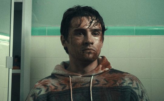 A man sitting in a green tiled bathroom centre frame, staring vacantly into the distance. He is covered in dirt, sweat and blood.