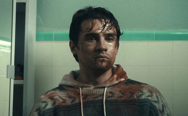 A man sitting in a green tiled bathroom centre frame, staring vacantly into the distance. He is covered in dirt, sweat and blood.