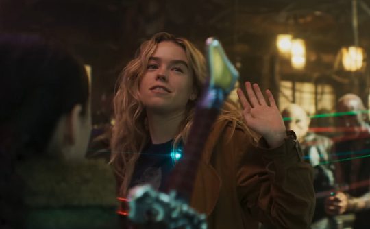 Supergirl stands center frame, her hands in the air, a cocky expression on her face. Multiple men surround her with weapons aimed at her.