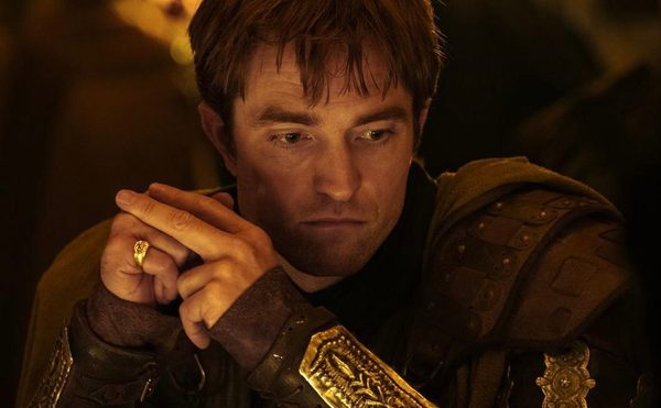 A man stares down, focused. He has light brown scruffy straight hair. he wears gold and brown armour.