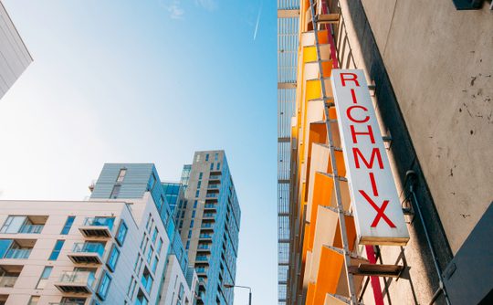 Upward view of colourful modern buildings beneath a bright blue sky, featuring a vertical “RICHMIX” sign; vibrant urban atmosphere.
