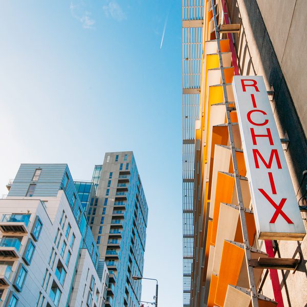 Upward view of colourful modern buildings beneath a bright blue sky, featuring a vertical “RICHMIX” sign; vibrant urban atmosphere.