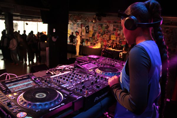 DJ wearing headphones performs on Pioneer decks and mixer in a colourful nightclub, with silhouetted dancers watching in the background.