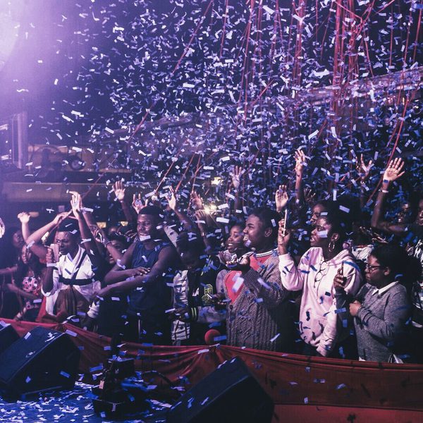 Performer on stage in a lively concert venue, surrounded by cheering crowd and falling confetti. Audience members wave hands, creating an energetic, celebratory atmosphere.