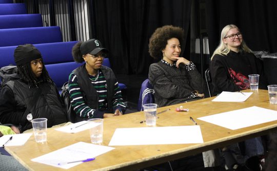 Four people sit at a wooden table in a relaxed, attentive manner, with papers and empty cups in front of them. The setting is a dimly lit room with blue seats in the background, conveying a casual and collaborative atmosphere.