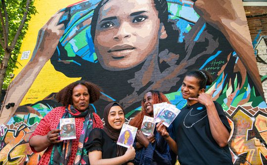 Four smiling people hold artwork in front of a vibrant mural depicting a person with a colourful headscarf, conveying a sense of community and celebration.