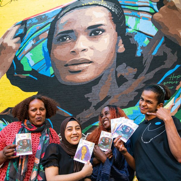 Four smiling people hold artwork in front of a vibrant mural depicting a person with a colourful headscarf, conveying a sense of community and celebration.