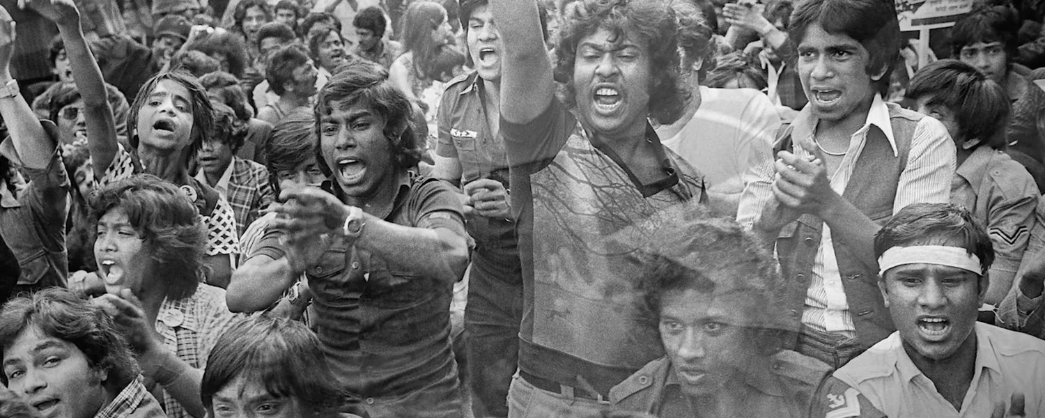 A black and white image of a large, passionate crowd of young people protesting. Some are shouting with raised fists, conveying energy and determination.