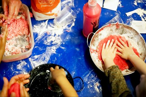 Children's hands kneading bright pink dough on a flour-dusted blue table. Flour package and red bottle visible, creating a playful, creative scene.