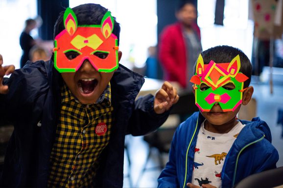 Two children wearing colorful animal masks smile and pose enthusiastically indoors. The mood is playful and vibrant, with a festive atmosphere.
