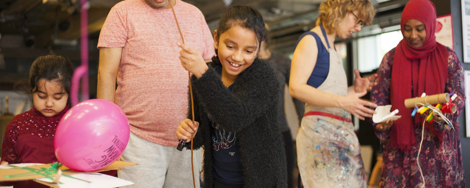 A cheerful young person holds a balloon and stick, surrounded by family and friends in a lively, colourful art workshop. Joyful, creative atmosphere.