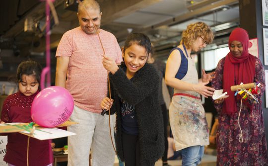 A cheerful young person holds a balloon and stick, surrounded by family and friends in a lively, colourful art workshop. Joyful, creative atmosphere.