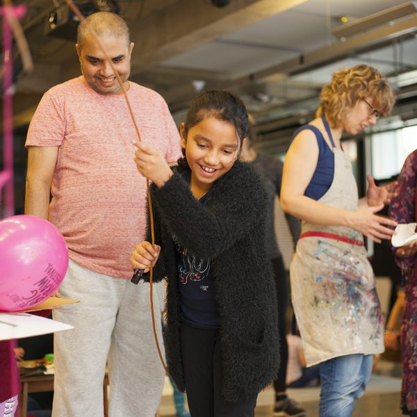 A cheerful young person holds a balloon and stick, surrounded by family and friends in a lively, colourful art workshop. Joyful, creative atmosphere.