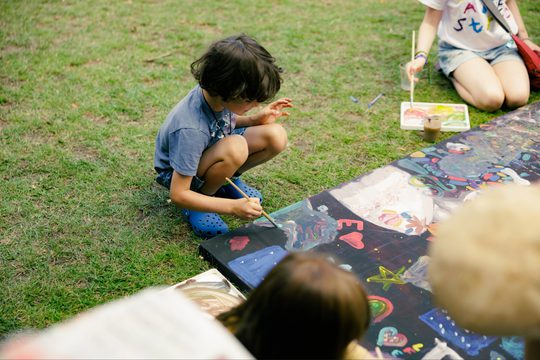 Child painting a colorful mural on the grass, surrounded by vibrant art supplies. The scene is lively and creative, emphasizing artistic expression.