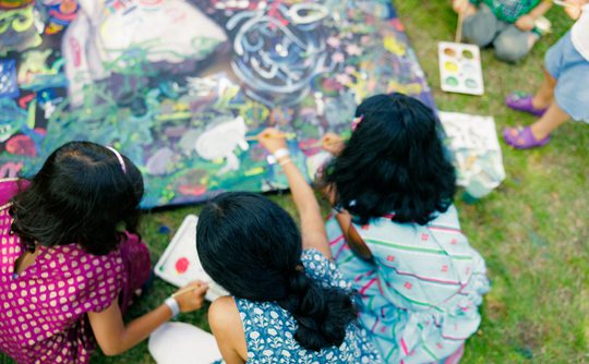 Three children are seated on grass, painting collaboratively on a colourful canvas. They are focused and engaged, surrounded by paint supplies.