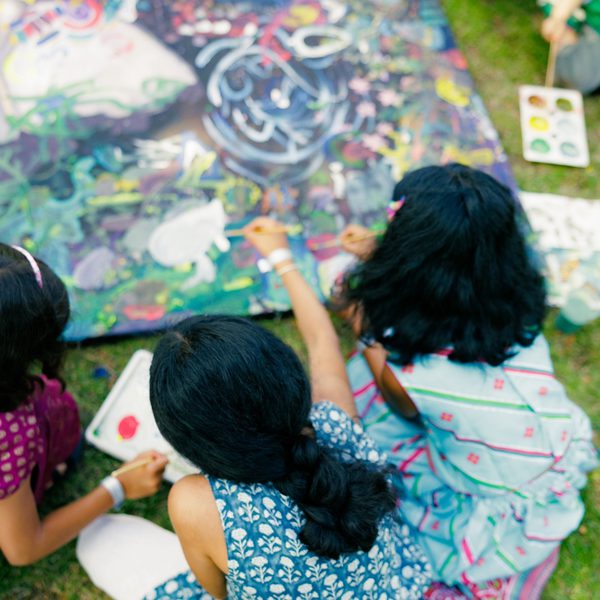 Three children are seated on grass, painting collaboratively on a colourful canvas. They are focused and engaged, surrounded by paint supplies.