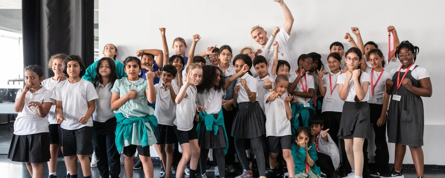 A diverse group of children and an adult strike confident poses with raised fists in a bright room. They wear casual attire and convey a sense of empowerment and unity.