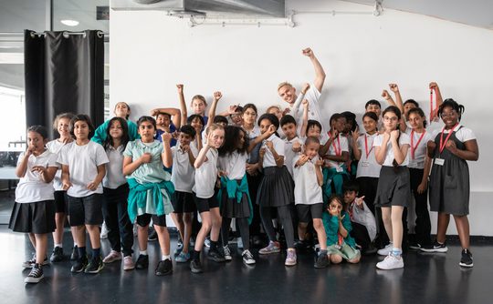 A diverse group of children and an adult strike confident poses with raised fists in a bright room. They wear casual attire and convey a sense of empowerment and unity.