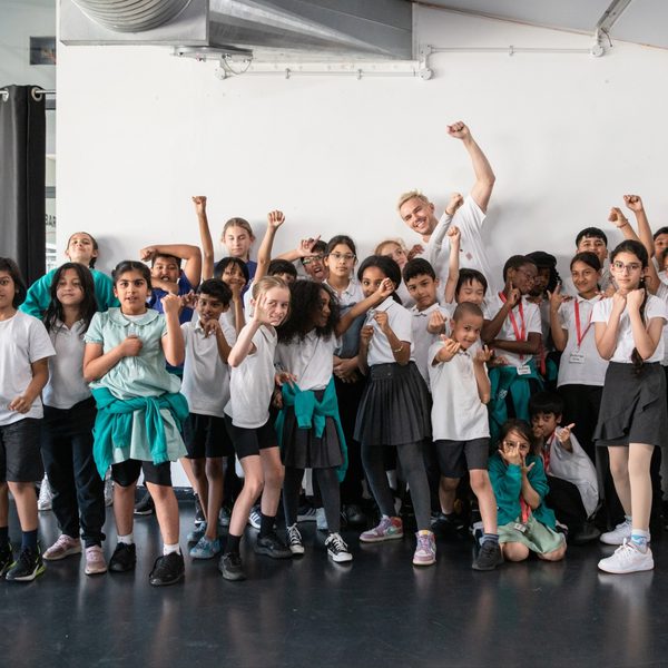 A diverse group of children and an adult strike confident poses with raised fists in a bright room. They wear casual attire and convey a sense of empowerment and unity.