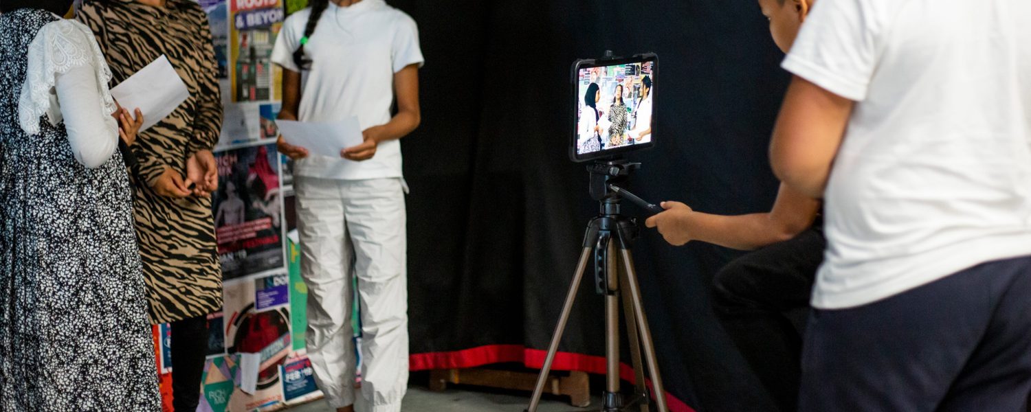 A group of young people, two in patterned dresses and one in a white outfit, stand before a camera placed on a tripod. They hold paper, suggesting a presentation or performance. The setting conveys a creative and engaged atmosphere.