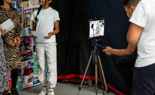 A group of young people, two in patterned dresses and one in a white outfit, stand before a camera placed on a tripod. They hold paper, suggesting a presentation or performance. The setting conveys a creative and engaged atmosphere.