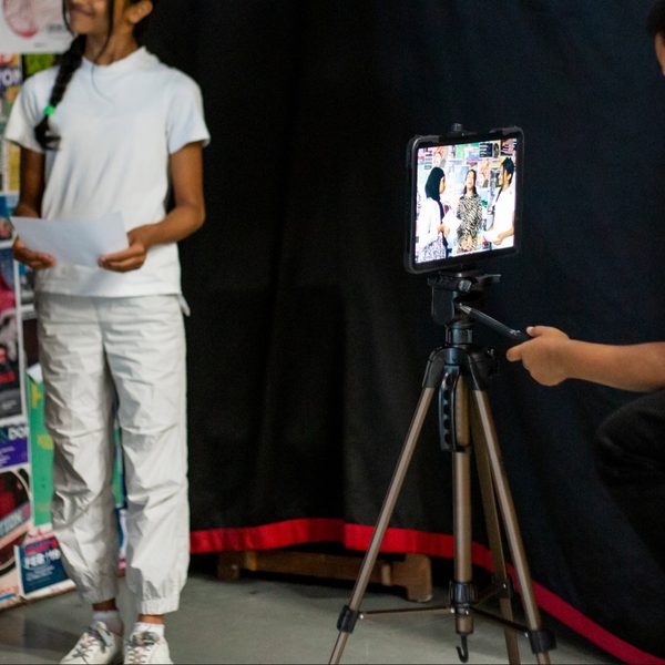 A group of young people, two in patterned dresses and one in a white outfit, stand before a camera placed on a tripod. They hold paper, suggesting a presentation or performance. The setting conveys a creative and engaged atmosphere.