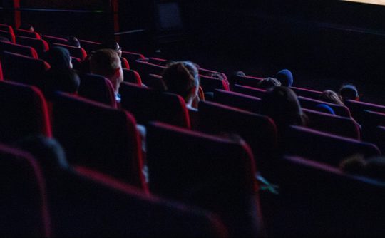 Dimly lit movie theatre with scattered audience in red seats, focused on a screen. The mood is calm, with anticipation and quiet enjoyment.