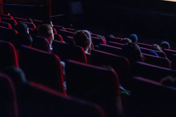 Dimly lit movie theatre with scattered audience in red seats, focused on a screen. The mood is calm, with anticipation and quiet enjoyment.