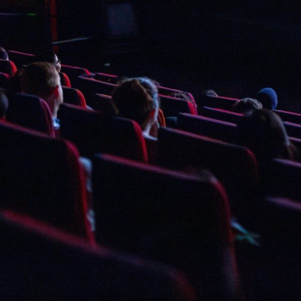 Dimly lit movie theatre with scattered audience in red seats, focused on a screen. The mood is calm, with anticipation and quiet enjoyment.