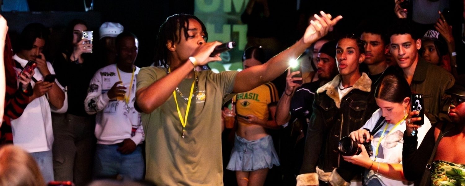 A lively performer in an olive shirt raps on stage, surrounded by an engaged crowd capturing the moment on phones. The atmosphere is energetic and vibrant.