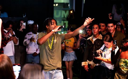 A lively performer in an olive shirt raps on stage, surrounded by an engaged crowd capturing the moment on phones. The atmosphere is energetic and vibrant.