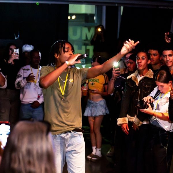 A lively performer in an olive shirt raps on stage, surrounded by an engaged crowd capturing the moment on phones. The atmosphere is energetic and vibrant.