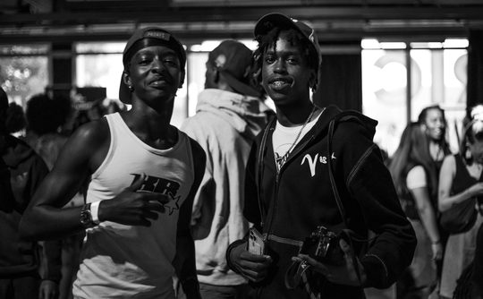 A black and white image of two smiling individuals in casual attire, standing in a crowded indoor setting. One holds a camera, conveying a lively atmosphere.