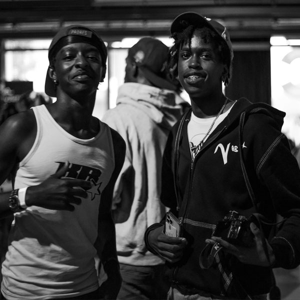A black and white image of two smiling individuals in casual attire, standing in a crowded indoor setting. One holds a camera, conveying a lively atmosphere.