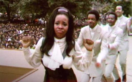 A woman dancing while staring at the camera, three men in white suits dancing behind her. Behind them is a massive crowd.