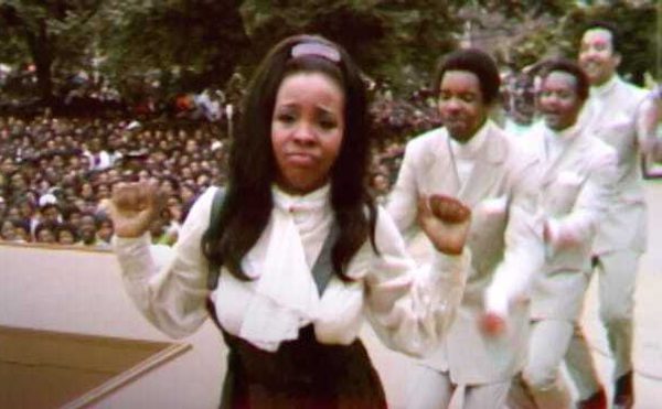 A woman dancing while staring at the camera, three men in white suits dancing behind her. Behind them is a massive crowd.