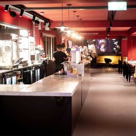 A modern bar with a long counter in a dimly lit space, featuring red walls and various equipment. Two people work behind the counter, creating a cozy, inviting atmosphere.