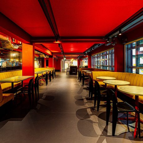 A modern interior with a vibrant red ceiling, yellow booths, and round tables. Large windows on the right create an inviting, warm atmosphere.