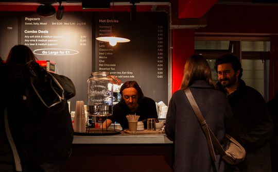 A cozy cafe scene with a barista preparing drinks behind the counter. Two customers wait, one smiling. Warm lighting and a menu above create a welcoming atmosphere.