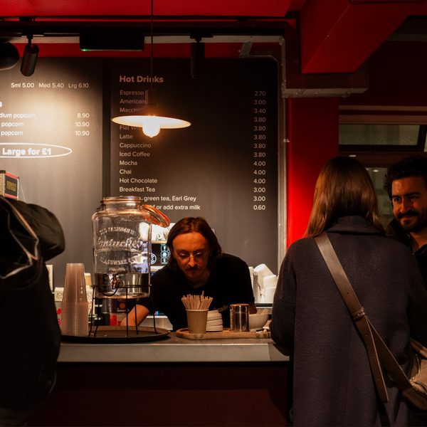 A cozy cafe scene with a barista preparing drinks behind the counter. Two customers wait, one smiling. Warm lighting and a menu above create a welcoming atmosphere.