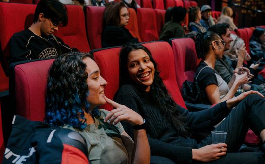 Two people sit in a theater, surrounded by others. They are smiling and chatting, creating a lively and relaxed atmosphere. Bright red seats fill the scene.