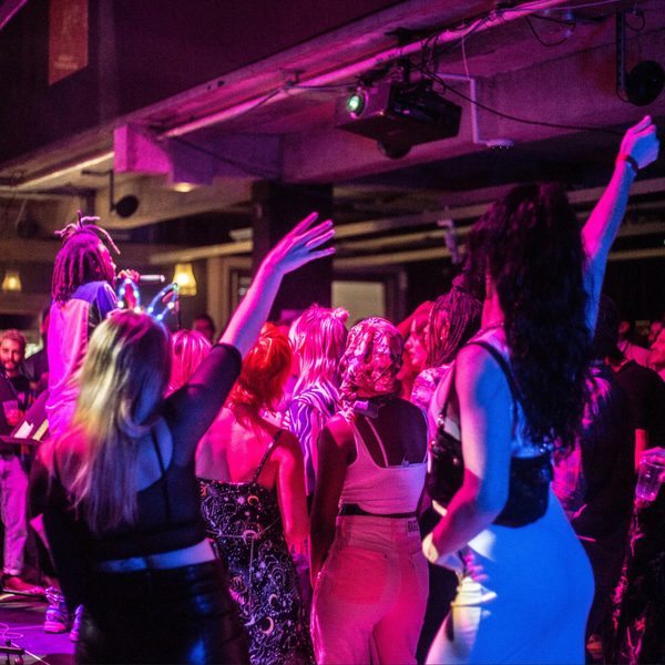 Vibrant nightclub scene with people dancing energetically under colorful purple and blue lights. A performer engages the lively crowd on stage.